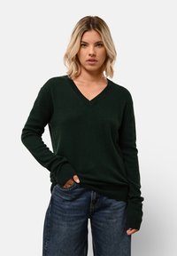Tmavě zelený svetr s výstřihem do V z měkkého materiálu, s dlouhými rukávy a žebrovanými manžetami, kombinovaný s volnými modrými džíny.