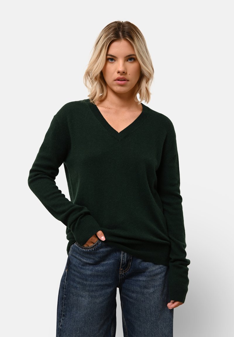 Tmavě zelený svetr s výstřihem do V z měkkého materiálu, s dlouhými rukávy a žebrovanými manžetami, kombinovaný s volnými modrými džíny.