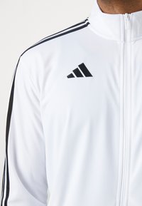 Hvid lynlås træningsjakke med sorte sidelister og Adidas-logo i sort på venstre bryst. Glat stof med en tætsiddende design.