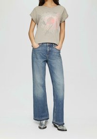 Beige grafisk t-shirt med en solstråledesign, tillsammans med vida ljusblå denimjeans och silverfärgade skor med spetsig tå.