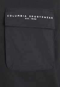 Sudadera negra con un bolsillo delantero bordado que presenta el texto "COLUMBIA SPORTSWEAR EST. 1938." La tela tiene un acabado suave y texturizado.