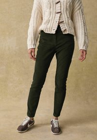 Donkergroene corduroy broek met een slim fit, gecombineerd met een crèmekleurige kabelgebreide trui en bruine sneakers. Geweven details en houten knopen.