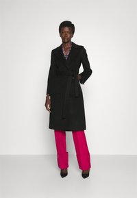 Schwarzer Trenchcoat mit Gürtel, großen Fronttaschen und geschlitzten Revers, kombiniert mit leuchtend pinkfarbenen, weit geschnittenen Hosen und schwarzen Absatzschuhen.