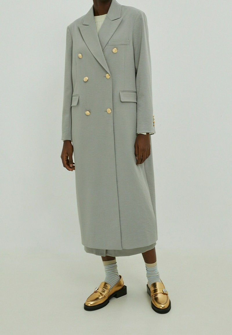 Manteau gris clair à double boutonnage avec des boutons dorés, deux poches avant et des manches longues. Associé à des mocassins dorés et des chaussettes rayées.