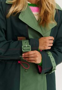 Zelený a tmavě modrý nepromokavý kabát s růžovým prošíváním, širokým límcem a nastavitelné manžety, obsahující stahovací prvek pro lepší přizpůsobení.