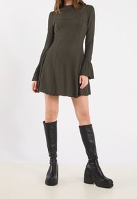 Kvinna i en mörk olivgrön långärmad miniklänning med utsvängda ärmar och svarta plattformskängor som når upp till knäna, mot en ensam bakgrund.