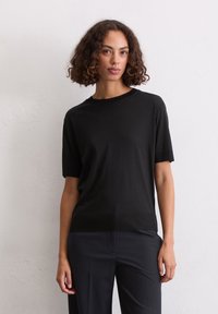 T-shirt nera a maniche corte realizzata in tessuto morbido, con scollatura girocollo classica, abbinata a pantaloni scuri su uno sfondo neutro.