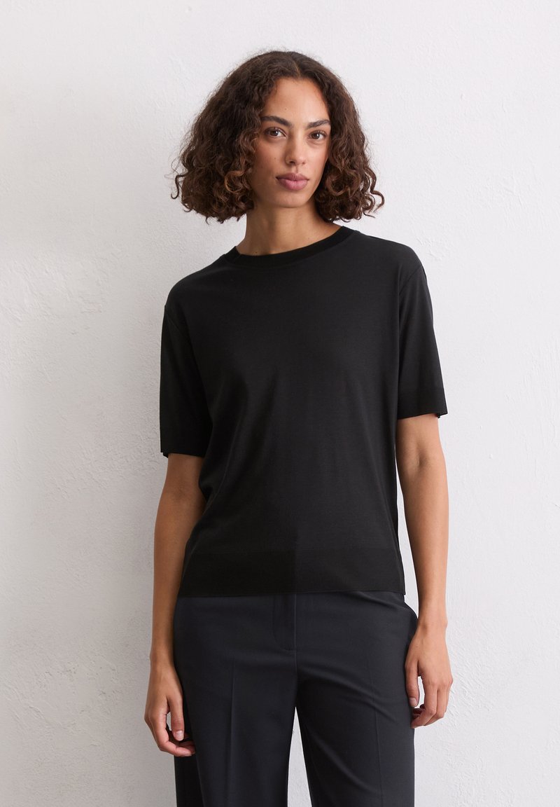 T-shirt nera a maniche corte realizzata in tessuto morbido, con scollatura girocollo classica, abbinata a pantaloni scuri su uno sfondo neutro.