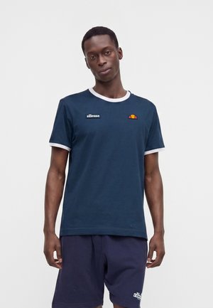 Uomo che indossa una t-shirt ellesse blu navy con rifiniture bianche e pantaloncini blu navy, in piedi davanti a uno sfondo bianco semplice.