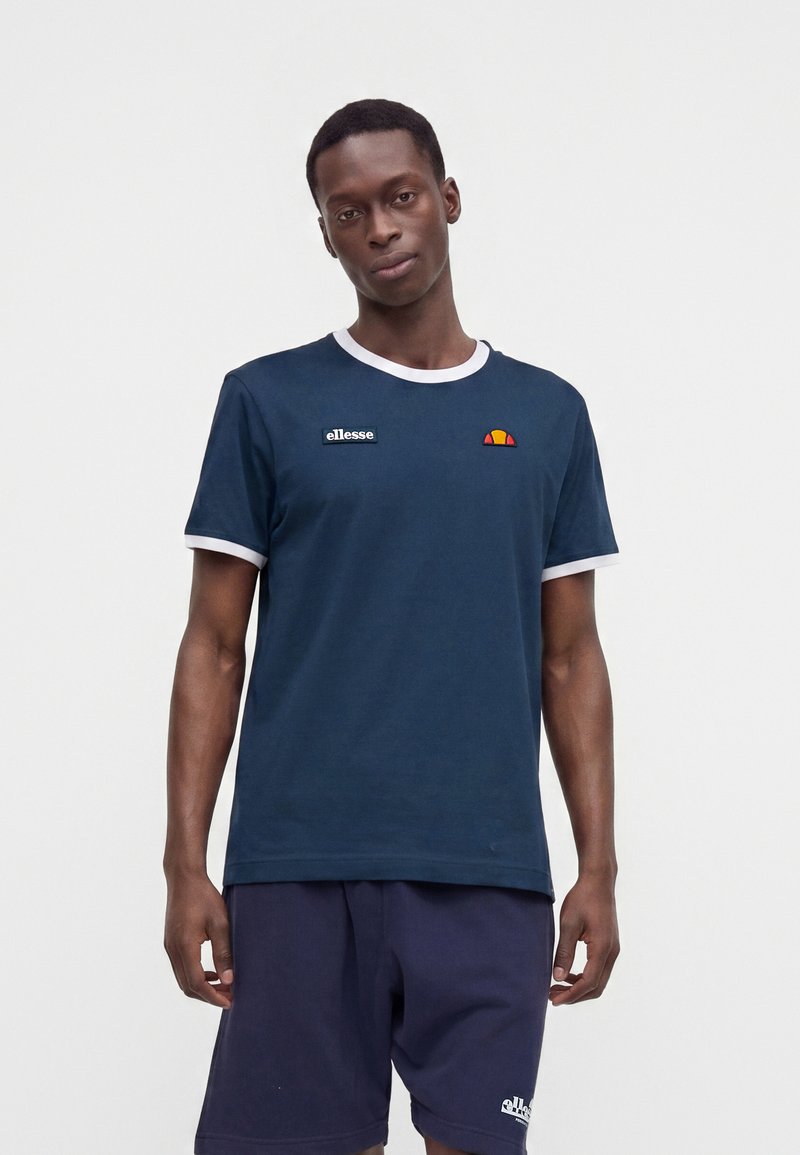 Homme portant un t-shirt ellesse bleu marine avec bordure blanche et un short bleu marine, debout devant un fond blanc uni.