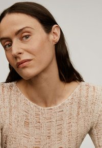 Jersey de punto rosa claro con un diseño calado. El modelo tiene el pelo castaño lacio y lleva puestos pequeños pendientes de oro. Fondo neutro.