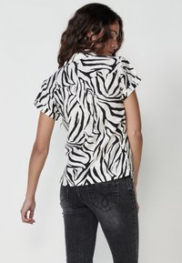 Camisa de manga corta con un estampado de cebra en blanco y negro, que presenta un cuello y una parte trasera con pliegues. Llevada con jeans gris oscuro.