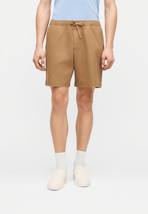 Mann trägt hellbraune Shorts mit Kordelzug, weiße Socken, beige Slipper und ein hellblaues Hemd, steht vor einem einfarbigen Hintergrund.