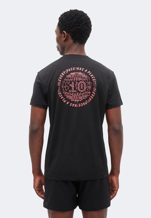 Mand, der vender væk, iført en sort T-shirt med et pink rundt mønster og teksten "EVERY SPACE HAS A PLACE" på ryggen, kombineret med sorte shorts.