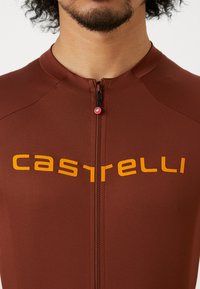 Bruin fietsoutfit met een kwart rits, voorzien van het "castelli"-logo in vet oranje over de borst. Effen, rekbare stof.