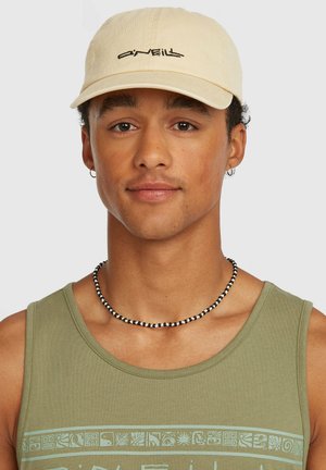 Gorra de algodón beige con logo negro bordado, usada por una persona con cabello castaño. La camiseta sin mangas verde presenta una banda gráfica azul claro y un collar de cuentas negras.