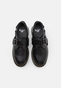 Scarpe in pelle nera con due cinturini spessi e fibbie. Design a punta tonda e cuciture gialle visibili lungo la suola. Suola in gomma antiscivolo.
