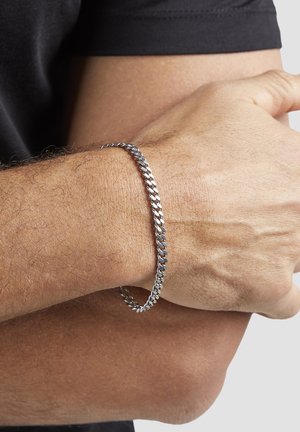 Nahaufnahme eines Handgelenks mit einem silbernen Kettenarmband, wobei eine Hand den gegenüberliegenden Arm umgreift.