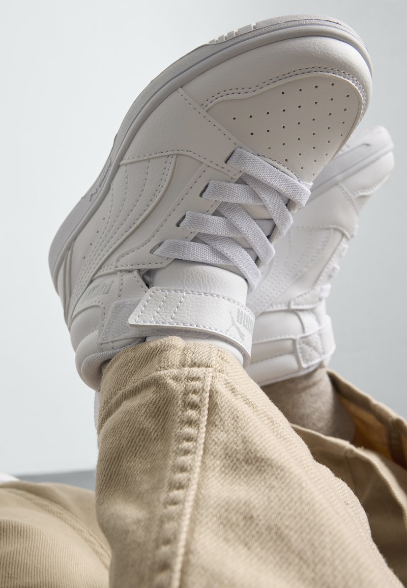 Witte leren sneakers met geperforeerde neus, platte veters en een klittenbandsluiting. Voorzien van subtiele branding en een rubberen zool. Draagt met tankleurige broek.
