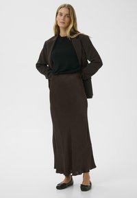 Long brown skirt with a smooth texture, accompanied by a black top and dark blazer, paired with flat black shoes. Simple, streamlined design.