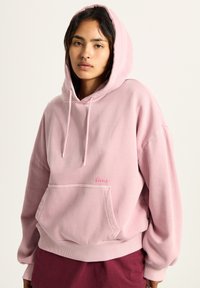 Pink hooded sweatshirt with a front pocket, relaxed fit, ribbed hem, and drawstring hood. Small logo embroidered in pink on the chest.