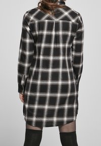 Abito camicia a quadri in bianco e nero con maniche lunghe, polsini con bottoni e una piega nella parte posteriore, presenta un orlo dritto e spacchi laterali.