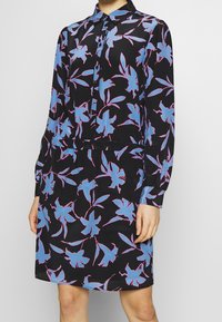 Abito con maniche lunghe, colletto abbottonato, realizzato in un tessuto nero decorato con motivi floreali rosa e blu. Mano posizionata sull'anca.
