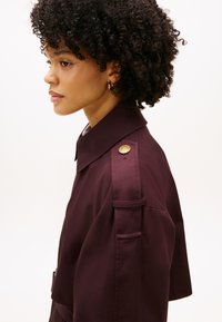 Μπορντό κοντό μπουφάν με δομημένο γιακά, λουράκια στους ώμους και λεπτομέρειες με χρυσά κουμπιά. Ομαλή υφή, με πλαϊνές τσέπες και ραμμένο σχέδιο.