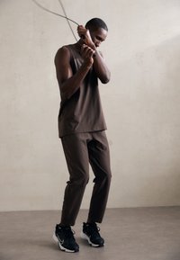 Haut sans manches marron foncé et pantalon de jogging assorti, associés à des chaussures de sport noires et blanches. Le modèle saute à la corde.