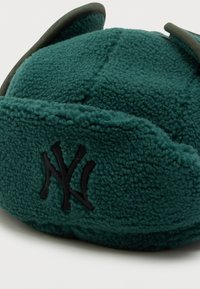 Zelená fleecová čiapka s ušnými klapkami, s čiernym vyšívaným logom. Materiál je mäkký a textúrovaný, navrhnutý pre teplo a pohodlie.