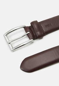 Cinturón de cuero marrón con hebilla de metal plateado, que presenta detalles de costura y el logo "POLO" en relieve en la correa. Textura suave en general.