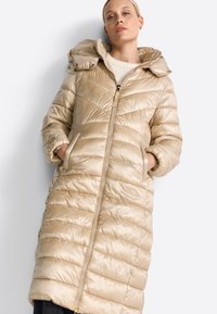 Beige pufferjas met capuchon, voorzien van een doorgestikt patroon, een ritssluiting aan de voorkant, en zijzakken. Gladde, glanzende stof met gouden accenten.