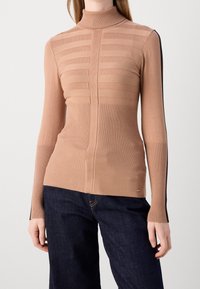 Beige coltrui met horizontale streepdetaillering, geribbelde textuur en lange mouwen met contrasterende zwarte accenten langs de zijkanten.