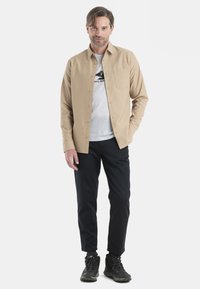 Camicia beige con bottoni e taschino sul petto, indossata sopra una maglietta grigia con grafica, abbinata a pantaloni neri e scarpe da trekking nere.