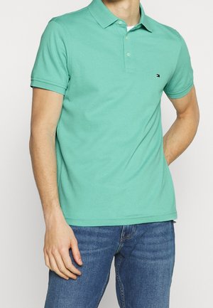 Homme portant un polo vert menthe à manches courtes avec deux boutons, petit logo sur la poitrine, et un jean bleu, debout avec une main dans la poche.