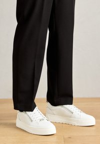 White leather sneakers with a textured finish, featuring a front zipper, flat laces, and a thick rubber sole, worn with black trousers.