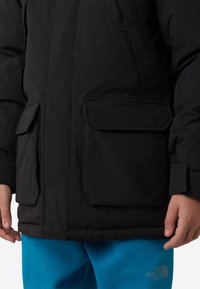 Μαύρο μπουφάν με μόνωση, με δύο τσέπες με καπάκι και κλείσιμο με φερμουάρ. Το υλικό είναι λείο με ματ φινίρισμα. Φορώντας με μπλε φόρμα.