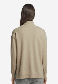 Frau mit langen, dunklen Haaren, die einen beigen Rollkragenpullover und hellgraue Jeans trägt, steht mit dem Rücken zu einem weißen Hintergrund.