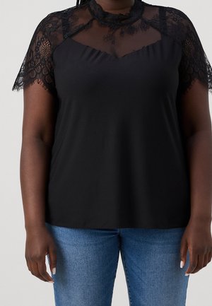 Frau trägt schwarzen Oberteil mit transparentem Spitzenjoche und kurzen Spitzenärmel, kombiniert mit blauer Jeans, steht vor neutralem Hintergrund.