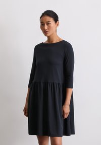 Mustmustere must dress, mille kolmeveerandi varrukatega, ümmarguse kaelusega ja kogutud seelikuosa. Omab lõtva istuvust ja siledat tekstuuri. Minimalistlik disain.