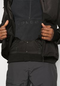 Personne portant une veste et un pantalon foncés, tenant la veste ouverte pour révéler un sweat-shirt en polaire noir avec le logo texte "VOLCOM".
