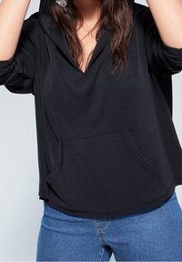 Nainen, jolla on päällään löysä musta huppari etutaskulla ja käärityt hihat, yhdistettynä sinisiin farkkuihin, näkyy keskivartalon osalta, eikä kasvoja näytetä.