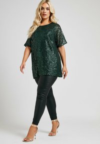 Top verde scintillante in paillettes con maniche corte e vestibilità ampia, abbinato a leggings neri aderenti e décolleté dorate con cinturini.