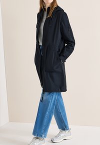 Femme portant une longue veste à capuche bleu marine, un pull gris, un jean large bleu et des baskets argentées, debout devant un fond uni.
