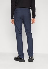 Pantalones azul marino con un corte ajustado, que cuentan con bolsillos traseros con cierres de botón y una textura suave, combinados con zapatos negros.