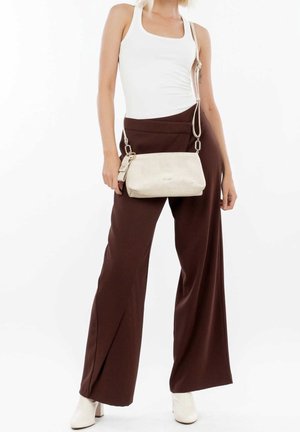 Mujer con top blanco sin mangas, pantalones marrones de talle alto y pernera ancha, botas blancas y llevando un bolso bandolera beige.