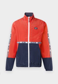 Punane ja tumesinine tuulejakk, millel on esimesel külgedel tõmblukk, seisev krae ja Reebok'i logo. Kätistel on triibuline detail.