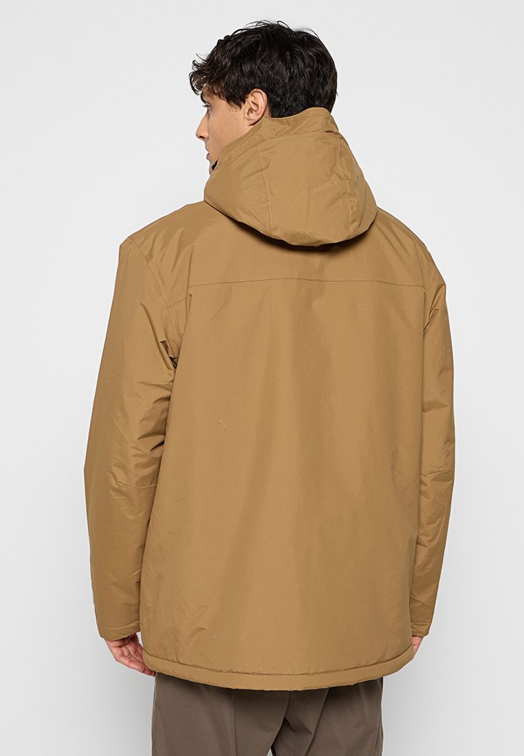 Homme portant une veste d'hiver à capuche marron, vu de dos contre un fond clair uni.