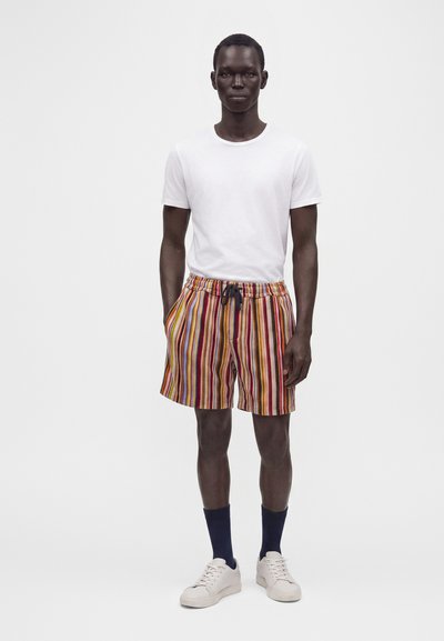 Homme debout portant un t-shirt blanc uni, un short rayé vertical multicolore, des chaussettes foncées et des baskets blanches sur un fond uni.