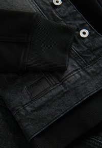 Dettaglio della giacca in denim nera con tessuto testurizzato, polsini a coste, bottoni a pressione argentati e un'etichetta nera con la scritta "Ombre" in rilievo sull'orlo.